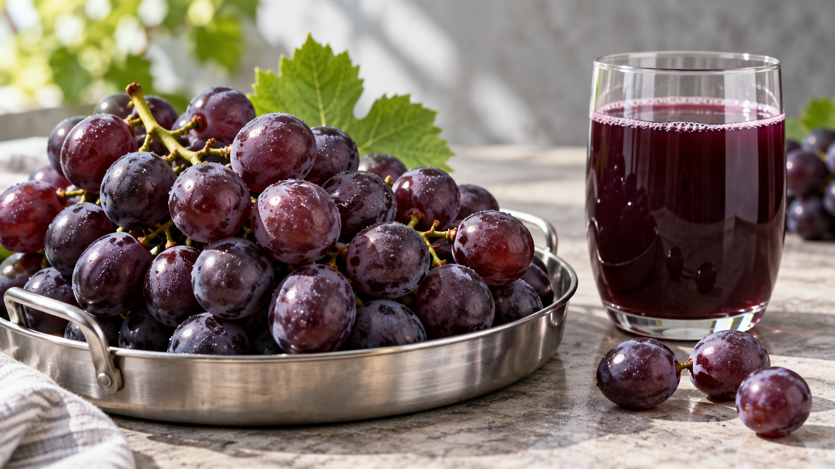 Close-up of purple grapes in a shallow steel tray beside a glass of dark grape juice on a clean stone surface, soft directional daylight from the side, shallow depth of field, realistic commercial food photography, landscape composition, no branding